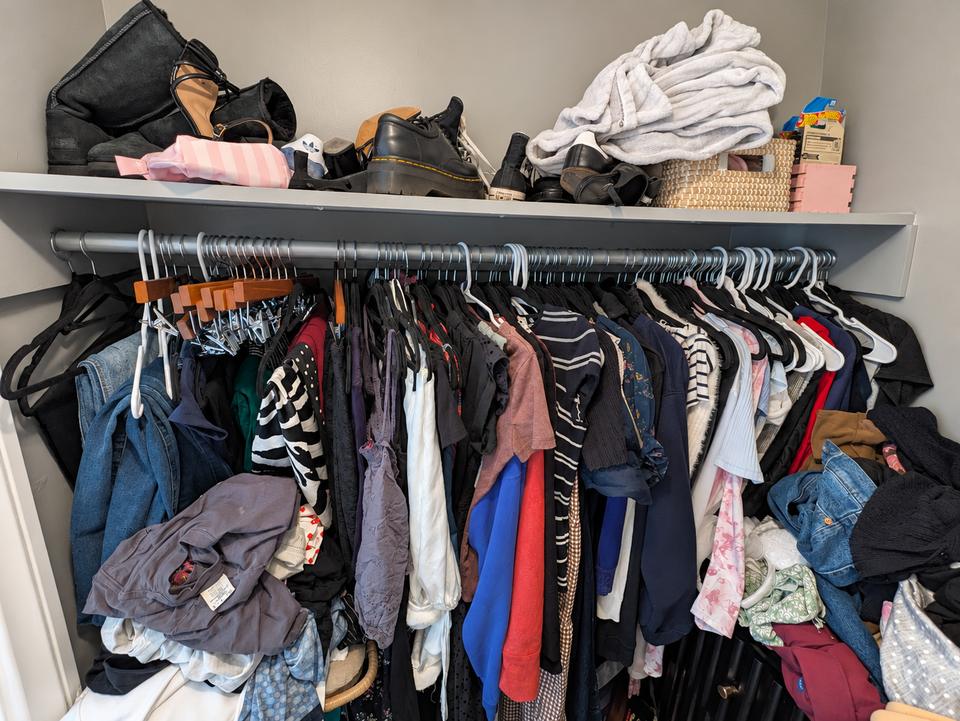 Closet before organizing, with crowded hanging clothes and belongings piled on the upper shelf