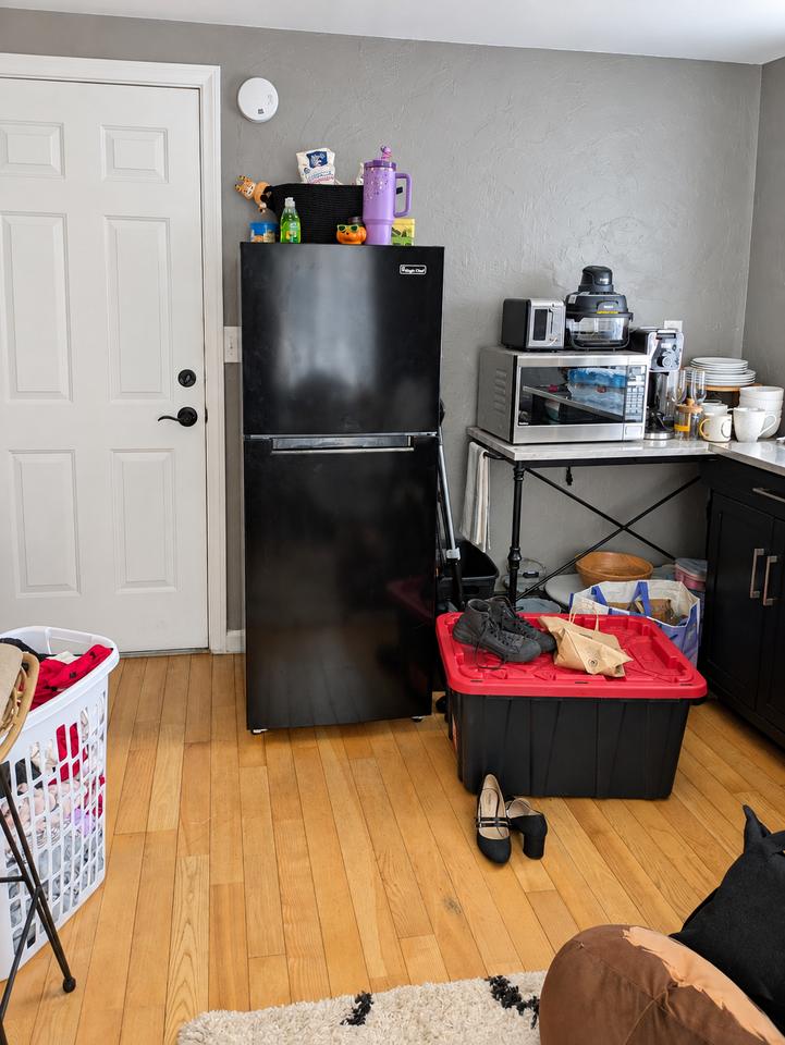 Studio kitchenette before organizing, with bins and belongings around the refrigerator and appliance table